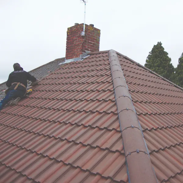 A roofer working on a brown tiled hip roof beside a brick chimney with lead flashing